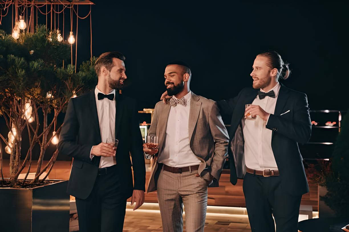 Three groomsmen in formal suits walking and laughing while holding whiskey glasses; a celebration highlighting personalized groomsmen gifts and custom engraved whiskey decanter sets.
